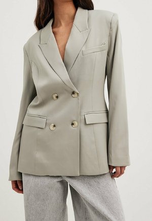 Kvinde iført en lysegrå dobbeltradet blazer med brede revers og matchende knapper, kombineret med lysegrå jeans.