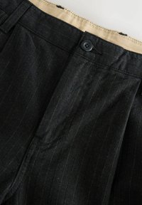 Schwarze Nadelstreifen-Hosen mit Knopfverschluss und beiger Innenbund, nah aufgenommen auf einer hellen Oberfläche.