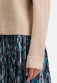 Beige gerippter Strickpullover mit einem Detail am Saum, kombiniert mit einem gefalteten Rock in Grüntönen, Blau und Schwarz.