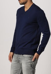 Granatowy sweter w serek wykonany z miękkiego materiału, z ribbowanymi mankietami i dołem, noszony z jasnoszarymi postarzanymi dżinsami.