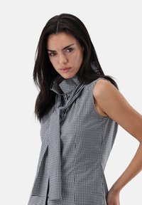 Sleeveless blouse in black and white gingham check pattern, featuring a high collared neckline with a tied accent and a fitted silhouette.