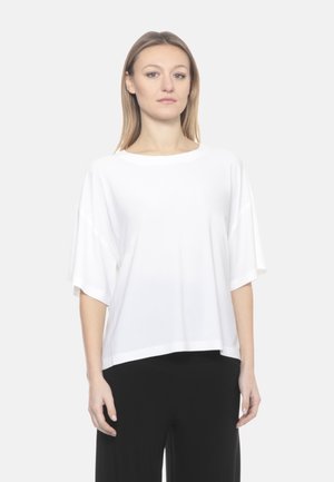 Giovane donna che indossa una maglietta bianca a maniche corte e pantaloni neri, in piedi davanti a uno sfondo grigio chiaro uniforme.