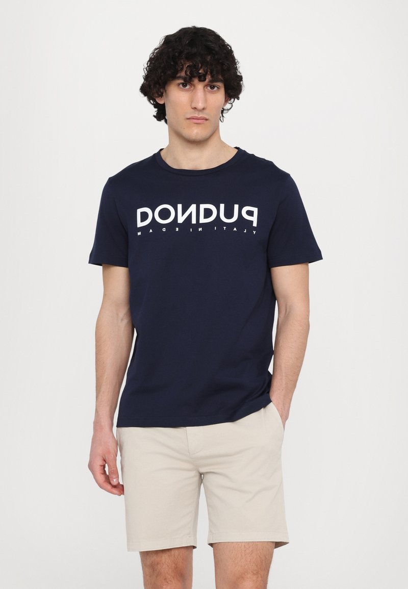 T-shirt blu navy in cotone, con scollo a girocollo, caratterizzata da un grande testo grafico bianco "PUDON" sul davanti. Abbinata a pantaloni corti beige.