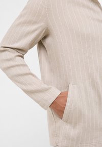 Top in lino beige con righe verticali bianche, dotato di maniche lunghe e tasche laterali. Texture morbida con un design dalla vestibilità comoda.