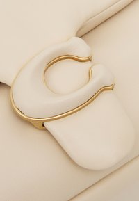 Off-white läderhandväska med detaljer som en unik loopad lås i guld och en slät yta på en semi-strukturerad design.