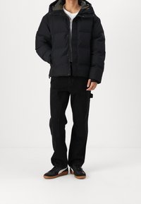 Mand iført sort dunjakke, sorte cargobukser, hvid t-shirt og sorte sneakers med brune såler, stående på hvidt gulv.