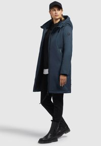 Gepolsterte marineblaue Parka mit Kapuze, Reißverschluss, Seitentaschen und glatter Textur, getragen über einem schwarzen Oberteil und schwarzen Hosen, kombiniert mit schwarzen Stiefeln.