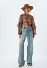 Braune Paisley-Bluse mit Rüschensäumen und gebundenem Kragen, kombiniert mit hellblauen weit geschnittenen Jeans und einem braunen, lässigen Hut.