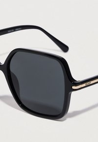 Čierne slnečné okuliare s obdĺžnikovým rámom, tmavými šošovkami a zlatými detailmi na nožičkách, opreté o svetlé pozadie.