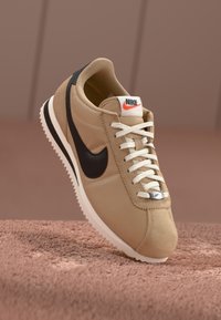 Nike sportbačiai smėlio ir juodos spalvos, turintys tekstilinį viršų, baltus raiščius, guminį padas ir retro dizainą su matomomis siūlėmis.