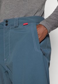 Ruka u džepu plavih hlača s bijelim šavom, nosi se s sivom košuljom dugih rukava, prikazuje zakopčani pojas i mali crveni oznaku.