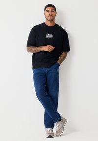 Camiseta negra de gran tamaño con gráfico en el pecho, vaqueros azules de mezclilla y zapatillas beige con detalles en gris claro. Posición de pie contra un fondo blanco.