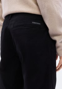 Κοντινό πλάνο ατόμου που φοράει μαύρο παντελόνι Calvin Klein και μπεζ πουλόβερ, εμφανίζοντας την πίσω τσέπη και τη λεπτομέρεια με την ετικέτα της μάρκας.