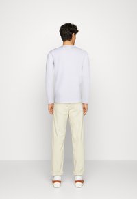 Camicia bianca a maniche lunghe, vista posteriore, abbinata a pantaloni beige chiaro. La camicia ha un collo rotondo e i pantaloni sono risvoltati alla caviglia.