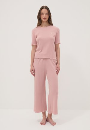 Donna in piedi scalza indossa una maglietta rosa chiaro a maniche corte larga e pantaloni ampi coordinati contro uno sfondo bianco semplice.