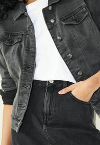 Osoba v černé džínové bundě přes bílé tričko, se svou pravou rukou v kapse černých džínů.