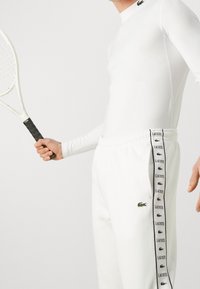 Bílé sportovní tričko s dlouhým rukávem a bílé tepláky s černými bočními pruhy, na kterých je nápis "LACOSTE"; obsahuje zelené logo krokodýla.