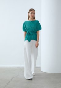 Blusa pieghettata color teal con vita cintata, maniche corte, abbinata a pantaloni bianchi a gamba larga e scarpe grigie con tacco. Design semplice e moderno.