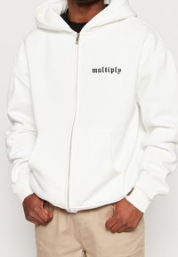 Witte rits-hoodie van zacht materiaal, met een voorkantzak en zwarte "multiply" tekst op de linkerkant van de borst.
