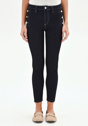 Femme portant un jean skinny bleu marine foncé avec des boutons dorés sur les poches de hanches et des mocassins beiges, debout devant un fond blanc.