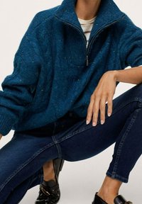 Suéter de punto azul con cuello alto y cremallera de cuarto. Textura acanalada con matices de color. Combinado con jeans ajustados oscuros y zapatos negros.