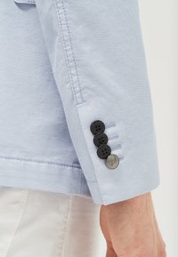 Světle modrý blazer s texturou, rukáv se čtyřmi černými knoflíky a jedním stříbrným knoflíkem, s kontrastním šitím a přizpůsobeným střihem.