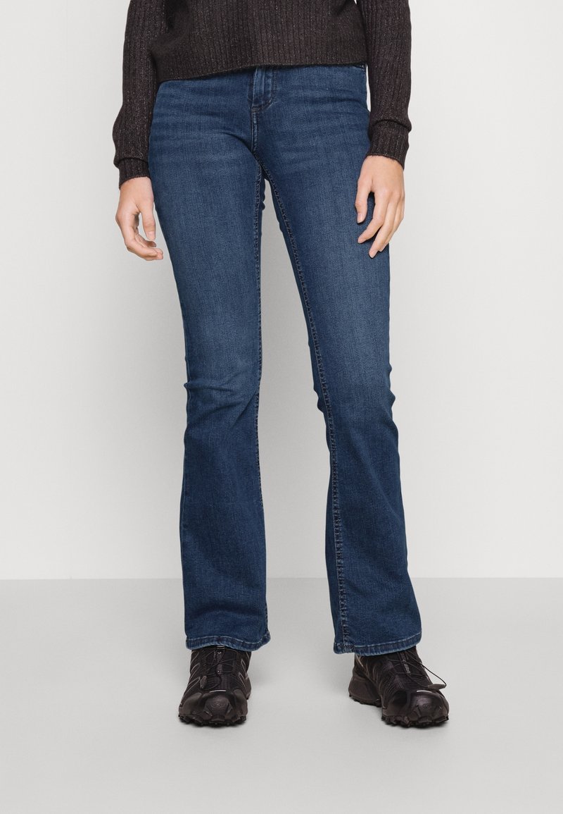 Personne portant un jean bleu foncé droit, des baskets noires et un pull noir à manches longues, posant devant un fond clair uni.