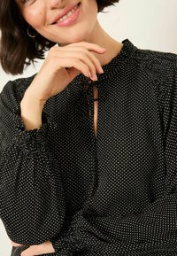 Schwarze Bluse mit weißen Punkten, hohen Ausschnitt und gerafften Manschetten. Verfügt über ein Schlüssel-Loch-Detail vorne und einen leichten Stoff.
