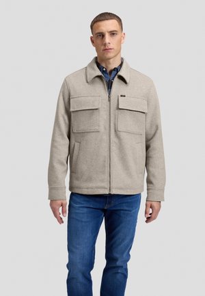 Jeune homme portant une veste beige zippée avec poches avant et un jean bleu, debout devant un fond gris clair uni.