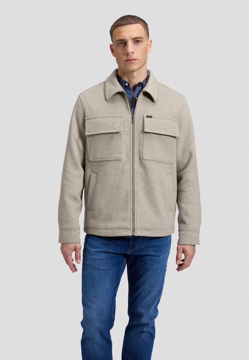 Jeune homme portant une veste beige zippée avec poches avant et un jean bleu, debout devant un fond gris clair uni.