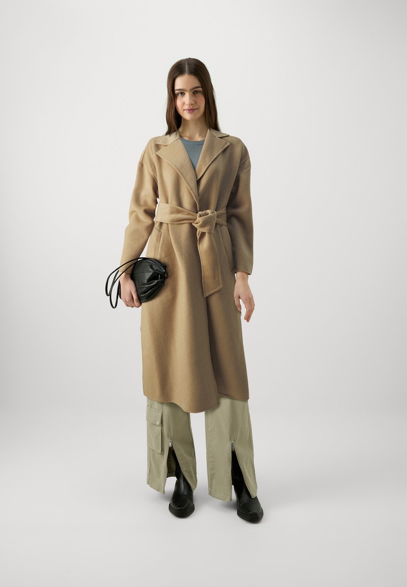 Beige Wollmischungs-Mantel mit tailliertem Gürtel, übergroßem Kragen, kombiniert mit hellgrünen Cargo-Hosen mit Reißverschlüssen und schwarzen Stiefeletten.