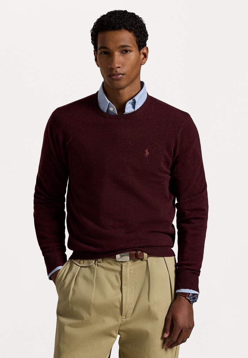 Suéter burdeos con cuello redondo y puños acanalados, superpuesto sobre una camisa de botones azul claro. Tejido de mezcla de algodón, acabado texturizado, combinado con pantalones caqui.