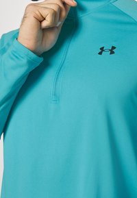 Türkisfarbener Sportpullover mit einem Viertelreißverschluss-Design, langen Ärmeln und einem schwarzen Logo auf der Brust. Glatte Stofftextur.