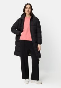 Schwarzer langer Steppmantel mit Kapuze, glänzendem Finish und Seitentaschen, getragen über einem rosa Hemd und schwarzen Hosen. Weiße Turnschuhe.