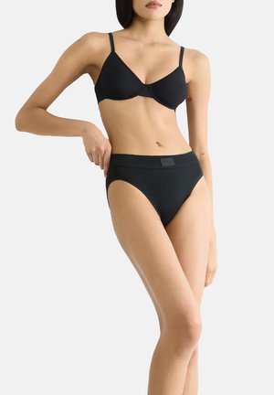 Frau trägt schwarzen BH und schwarze Unterwäsche mit hohem Bund, steht und richtet den Unterhosenbund vor einfarbigem Hintergrund.