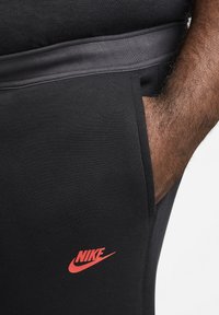 Schwarze Nike Sportanziehung mit rotem Logo, glatter Textur, schlankem Design und einer Seitentasche. Der Bund ist kontrastierend grau.