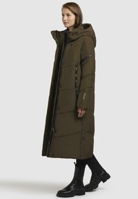 Dlouhý olivově zelený péřový kabát s kapucí, s předním zipem, na patentky a prošívaným designem. Nosené s černými kotníkovými botami.