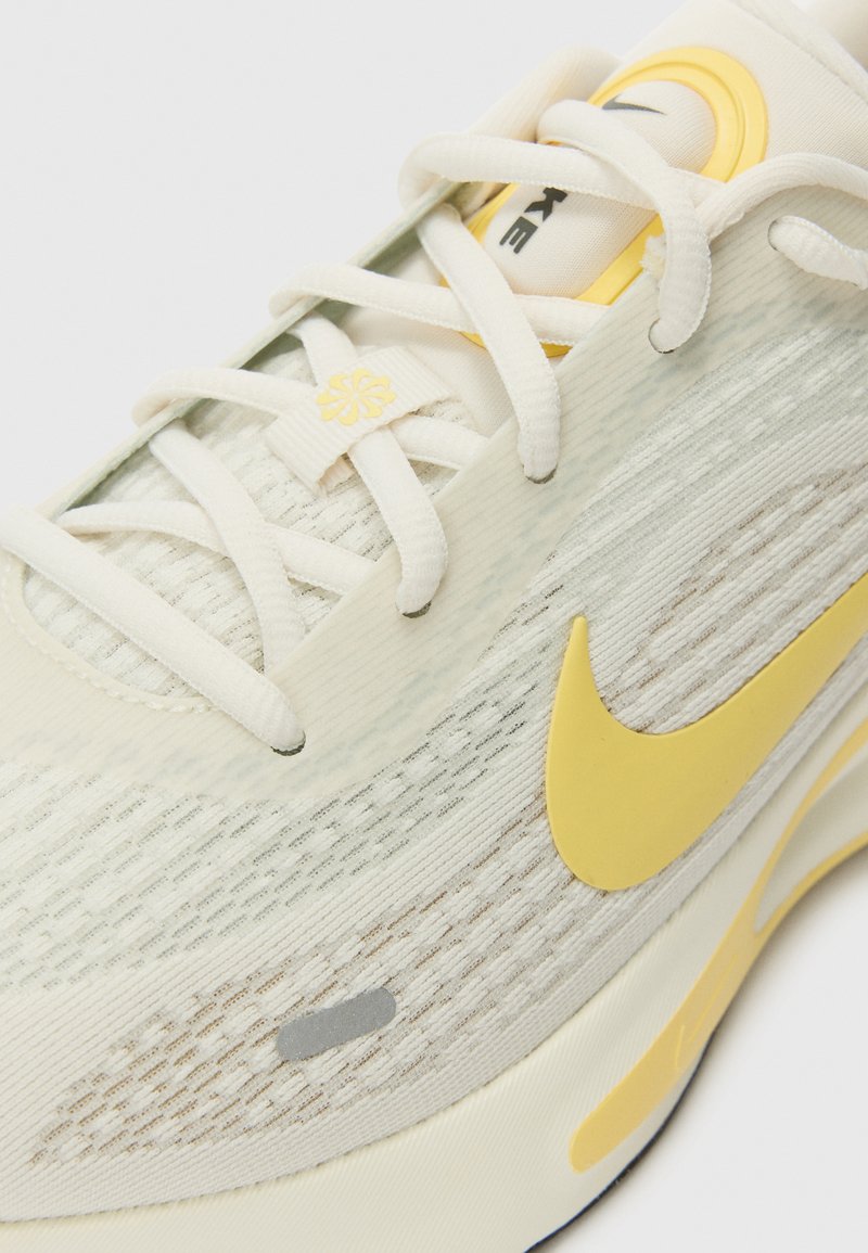 Baskets de sport beige clair avec tige en mesh texturé, accents jaunes et lacets blancs. Présente un swoosh distinctif de Nike et une semelle amortissante.