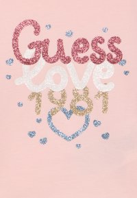 Γκλιτερ κείμενο «Guess love 1981» σε ροζ, λευκό και χρυσό σε ροζ φόντο, περιτριγυρισμένο από μικρές μπλε καρδιές με γκλίτερ και μια μεγάλη καρδιά.