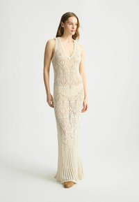 Abito lungo in maglia beige con scollatura a V profonda, caratterizzato da un corpetto fantasia e gonna plissettata. Realizzato in tessuto leggero e testurizzato.