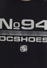 Svart T-shirt med djärva vita grafiker: "No 94" och "DC Shoes," med en logotyp och silverdetaljer. Klassisk rund halsringning och mjuk textur.