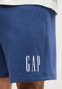 Μπλε casual σορτς με πλαϊνή τσέπη και λευκό λογότυπο "GAP", φορεμένα από άτομο που φοράει λευκό τοπ.