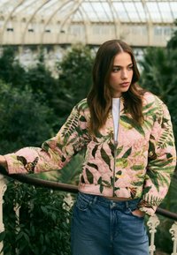 Gestepptes Bomberjacke in Pink mit botanischen Drucken, featuring grünen Blättern und Schmetterlingsakzenten, kombiniert mit blauen Jeans.