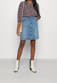 Femme portant un chemisier à manches longues fleuri, une jupe en jean boutonnée sur le devant, des bottines blanches et un petit sac à bandoulière beige, debout devant un fond uni.