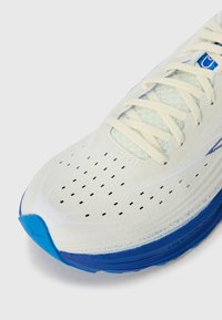 Chaussure de course avec un dessus crème clair, des accents bleus, un design perforé et une semelle texturée. Caractéristiques : lacets plats et capuchon de protection renforcé.