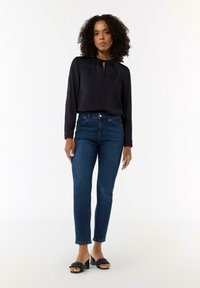 Granatowa bluzka z długim rękawem o zbieranym dekolcie, zestawiona z dopasowanymi niebieskimi dżinsami i czarnymi sandałami na obcasie o fakturowanej powierzchni.