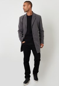 Grau Wolle Übermantel mit Reverskragen, zwei Knöpfen, kombiniert mit einem schwarzen strukturierten Pullover, schwarzen Hosen und schwarzen Sneakers.