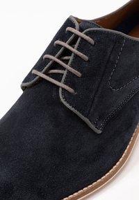 Chaussure en suède marine avec des lacets gris, bout rond, couture contrastante et semelle brun clair. Surface texturée et détails de design minimalistes.