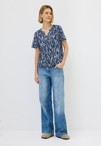 Mörkblå mönstrad kortärmad topp med V-hals, kombinerad med ljusblå vida jeans och beige skor, uppvisad mot en vit bakgrund.