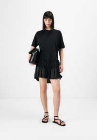 Zwarte jurk met een gelaagde ruffle zoom, korte mouwen en een losse pasvorm, gecombineerd met zwarte sandalen en een zwarte clutch. Gladde stoffen textuur.